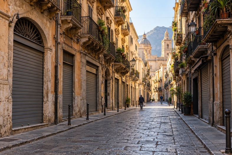 Strada ispirata al centro storico di Palermo al mattino presto, con numerose serrande abbassate e pochi passanti.
